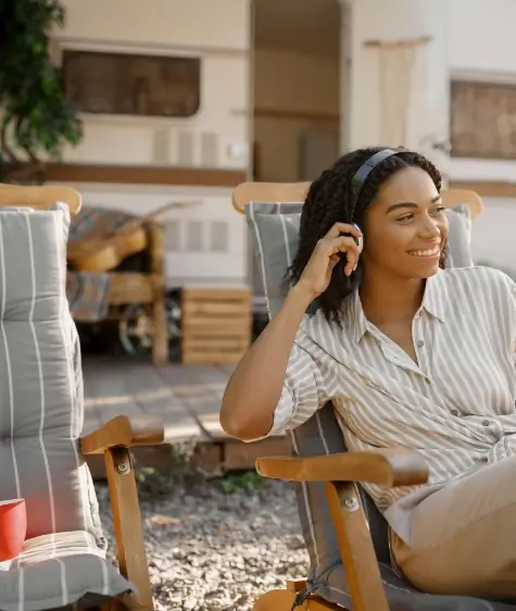 Woman relaxing in a chair outside an RV, smiling while using headphones.