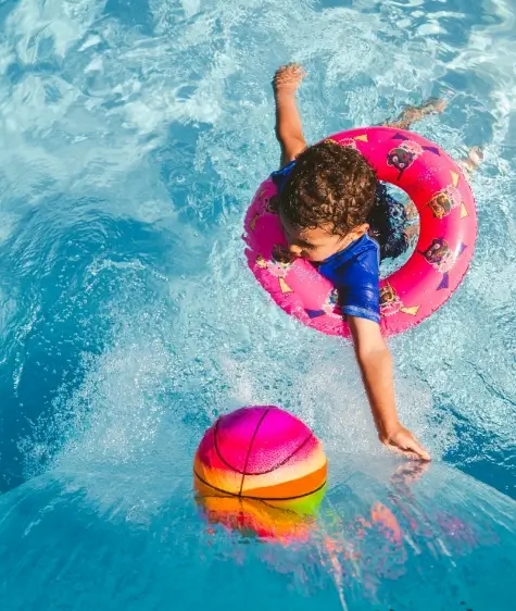 A child in an inflatable ring reaches for a basketball in a pool.